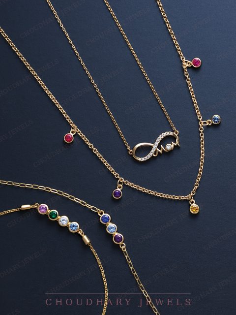 Yellow gold necklaces and bracelets on a navy background, including an infinity pendant with diamonds and the word Mom, a chain with colored gemstone drops and a bracelet with a row of colored stones.