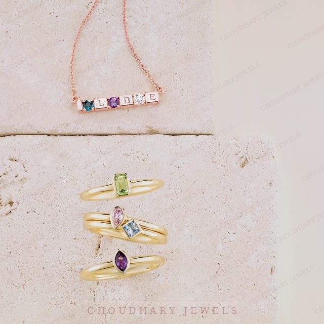 A rose gold bar necklace with letters and colored gemstones, above three stacked yellow gold rings with a green, a pink and blue, and a purple gemstone, on cream stone.