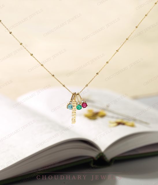 A yellow gold beaded chain necklace with small charms set with round blue, green and pink gemstones, above an open book.