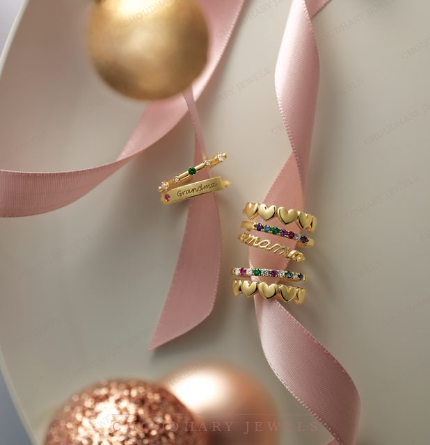 Yellow gold rings with rows of colored gemstones, hearts and the word mama, stacked on pink ribbon with rose gold ornaments.