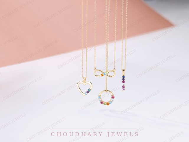 Four yellow gold pendant necklaces with colored gemstones: a heart, an infinity symbol, a circle and a vertical bar, hanging against a pink and white background.