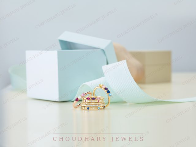 Yellow gold stacking rings set with rows of colored gemstones, beside an open light blue gift box and ribbon.