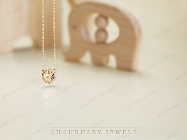 A yellow gold pendant necklace with a small round charm set with colored gemstones, beside a wooden toy elephant.