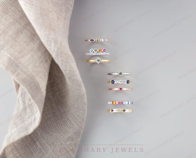 Seven yellow and white gold stacking rings set with small colored gemstones, beside a folded linen cloth on a gray background.