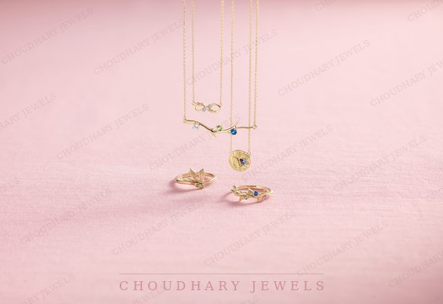 Yellow gold pendant necklaces and two rings with small gemstones, including an infinity pendant and a heart pendant, on a pink background.