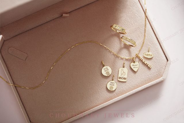 A yellow gold chain with engraved charms and tags, some set with small gemstones, displayed in a cream jewelry box.