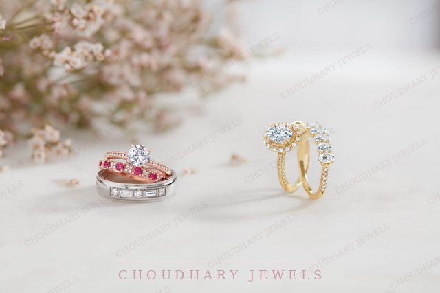 Two bridal ring sets on a white surface with dried flowers: a white gold diamond solitaire stacked with a pink stone band and a baguette diamond band, and a yellow gold halo engagement ring with matching diamond bands.