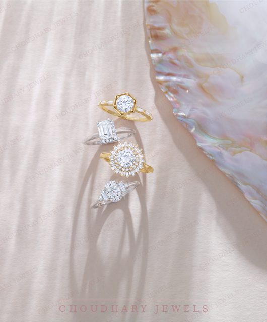 Four engagement rings on white linen beside an iridescent shell: a yellow gold hexagon halo, a white gold emerald-cut ring, a yellow gold round halo and a white gold kite-shaped ring.