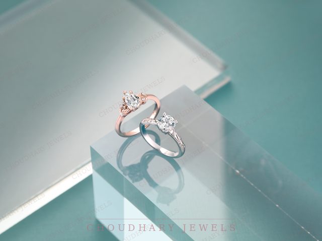 A rose gold three-stone engagement ring and a white gold round diamond ring on a clear acrylic block against a teal background.