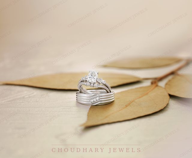 A white gold oval diamond engagement ring stacked with diamond wedding bands, resting on dried leaves.