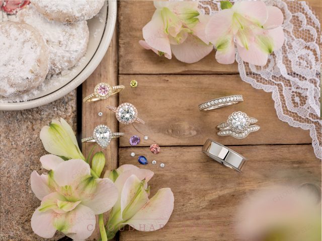 Engagement rings and bands on a wooden table with pink flowers and lace, including halo rings, a solitaire and loose colored gemstones.