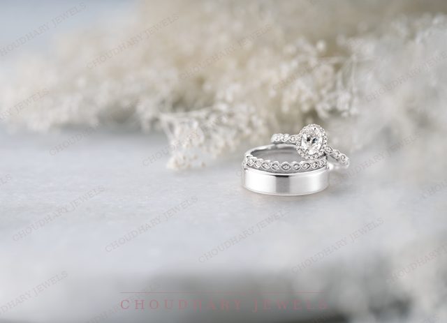 A white gold oval diamond engagement ring stacked with a diamond band beside a white metal men's band, on a pale blue surface.