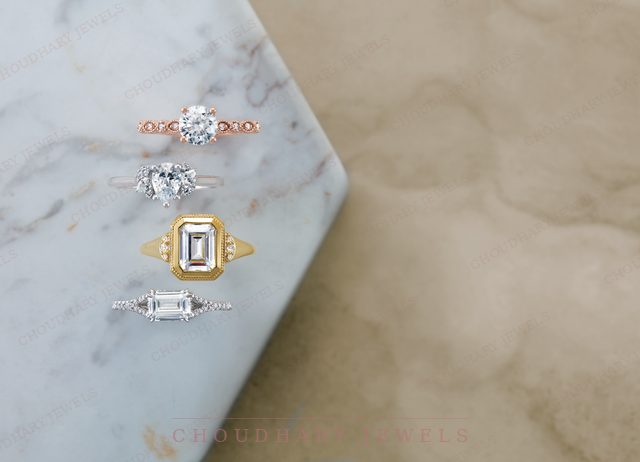 Four engagement rings on blue-veined marble: a rose gold round diamond with a beaded band, a white gold round diamond, a yellow gold emerald-cut halo and a white gold baguette ring.