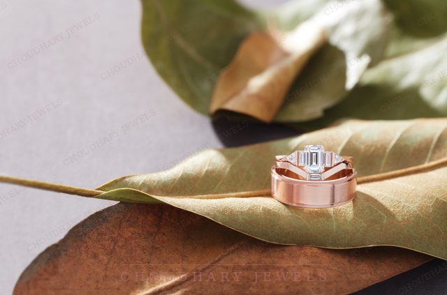 A rose gold engagement ring with an emerald-cut diamond and baguettes, resting on a large green leaf.