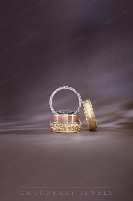 A stacked men's band set: a white metal band, a two-tone band with a rose and yellow engraved pattern, and a yellow gold band, on a purple background.