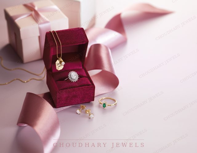 A burgundy ring box with a halo engagement ring and a gold pendant necklace, beside a pink gift box, pink ribbon and small gemstone rings.